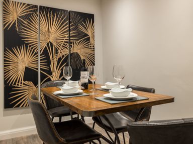 A dining table set with plates bowls wine glasses and a bottle of wine four dark chairs and a four panel palm leaf artwork on the wall at Ullswater View Apartment Ullswater