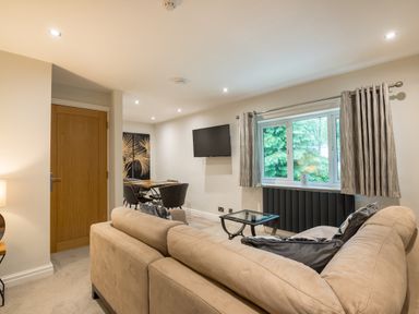 A living room with a beige sofa a glass coffee table wall-mounted television dining table and chairs near a window with curtains at Ullswater View Apartment in Ullswater