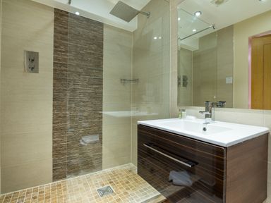 A bathroom with a walk-in shower, a dark wood vanity with a white sink, a large mirror, and beige tiled walls at Ullswater View Apartment in Ullswater