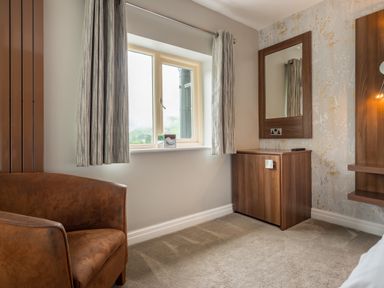A corner of a room with a brown armchair a window with curtains a wooden cabinet with a mirror above it and floral wallpaper at Ullswater View Apartment in Ullswater