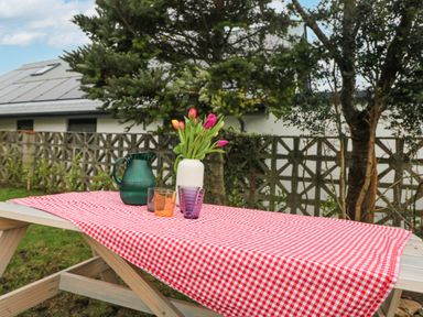 An outdoor wooden picnic table with a red and white checkered tablecloth a green jug purple and orange glasses and a white vase with tulips in a garden at Bod A Quinte in Carbis Bay