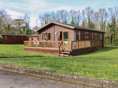 A wooden cabin with a railing deck and stairs surrounded by grass and trees at 3 The Glade in St Minver Rock