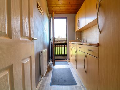 A narrow kitchen with wooden cabinets a sink two grey mats a white door and an open exterior door at 3 The Glade in St Minver Rock