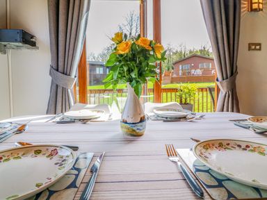 A dining table set with plates and cutlery a vase with yellow flowers on the table and a view of a wooden cabin outside through glass doors at 3 The Glade in St Minver Rock