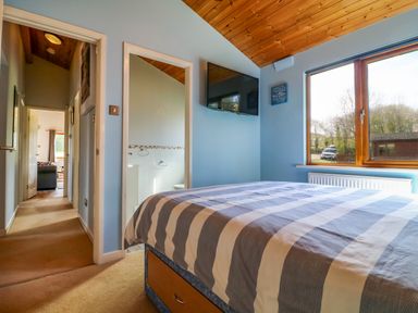 A bedroom with a bed striped bedding a wall-mounted tv window and an en suite bathroom at 3 The Glade in St Minver Rock