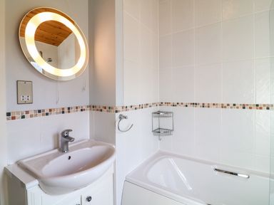 A bathroom with a sink, a lighted circular mirror, a towel ring, and a bathtub with a corner shower caddy at 3 The Glade in St Minver Rock