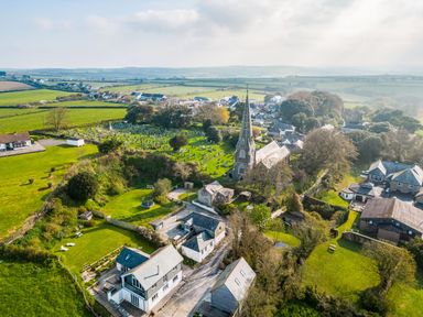 An aerial view of a village with a church and graveyard surrounded by houses and green fields at 3 The Glade in St Minver Rock