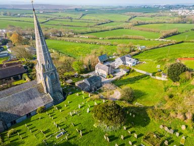 An aerial view of a stone church with a tall spire surrounded by a graveyard and green fields at 3 The Glade in St Minver Rock