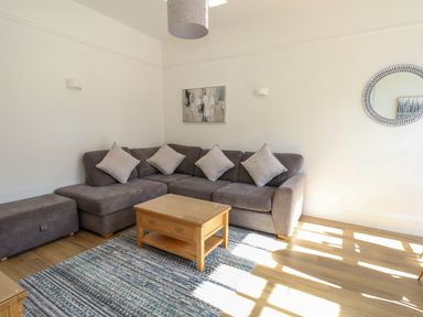 A living room with a grey sectional sofa and cushions a wooden coffee table a patterned rug a round mirror and wall art at Splash in Weymouth