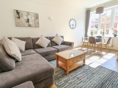 A living room with a grey sofa, a wooden coffee table, a glass dining table with chairs, and large windows at Splash in Weymouth