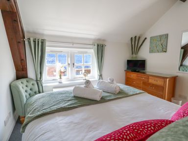 A bedroom with a bed with rolled towels on it a green chair a wooden dresser with a TV and a window with green curtains at Splash Weymouth