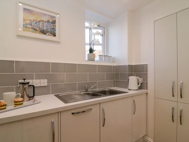 A kitchen with a double sink white cabinets a white electric kettle and a tray with a coffee press cup and muffins at Splash in Weymouth