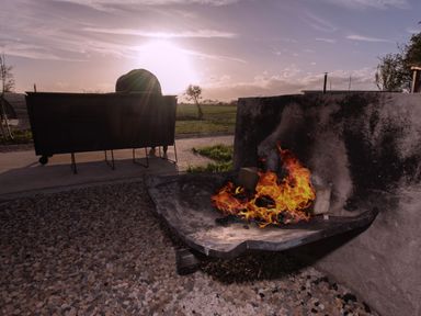 An outdoor fire pit with flames and a person sitting on a bench facing a sunset at Glamping Pod 2 Presence in Lebberston