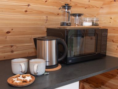 A kitchen counter with two mugs of tea a plate of star shaped cookies an electric kettle a microwave and glass containers on a wooden wall at Glamping Pod 3 Harmony Lebberston