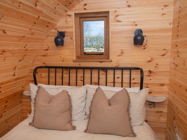 A bedroom with wooden walls a bed with beige pillows a small window and two black wall lamps at Glamping Pod 4 Truce Lebberston