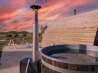An outdoor wooden hot tub with a metal chimney next to a wooden picnic table and a curved wooden cabin at Glamping Pod 4 Truce in Lebberston