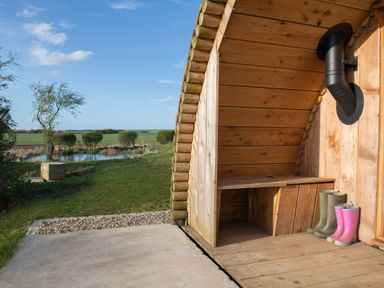 A wooden shelter with boots and a black stove pipe next to a grassy area with a pond at Glamping Pod 5 Shelter in Lebberston