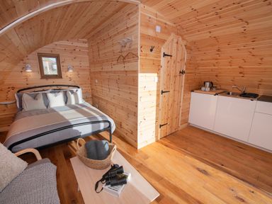 A wooden room with a bed pillows bedside lamps a small kitchen area a wooden table with a basket binoculars and books and a door at Glamping Pod 5 Shelter in Lebberston