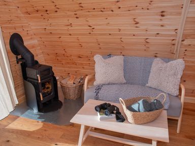 A wooden interior with a small stove and a basket of wood next to a sofa with cushions and a throw and a wooden table with a basket holding rolled towels and binoculars Glamping Pod 5 Shelter Lebberston