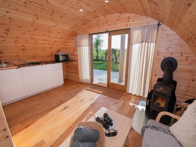 An interior view of a wooden pod with a kitchenette a wood burning stove glass doors with curtains and a small table with a basket and binoculars at Glamping Pod 5 Shelter in Lebberston