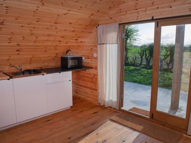 A small kitchen area with white cabinets a sink a microwave and glass door leading outside at Glamping Pod 5 Shelter Lebberston