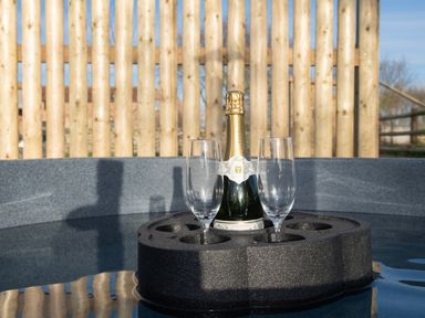 A bottle of champagne and two empty glasses on a floating holder in a hot tub with a wooden fence background at Glamping Pod 5 Shelter in Lebberston
