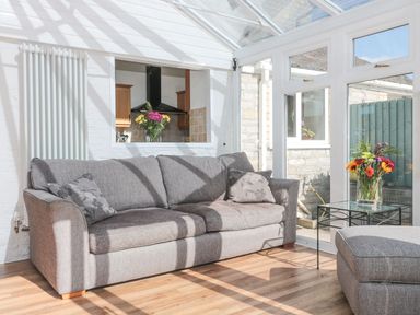 A conservatory with a gray sofa and cushions a glass table with flowers and wooden floor at Metcalfe House in Compton Dundon