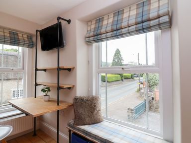 A corner workspace with a wall-mounted TV a wooden desk and shelves and a cushioned window bench at Metcalfe House in Compton Dundon