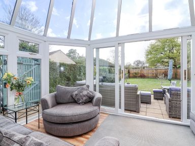 A conservatory with a grey armchair, a small glass table with flowers, and sliding glass doors opening to a garden with outdoor seating at Metcalfe House in Compton Dundon