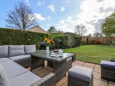An outdoor patio with a wicker table and sofa set with a vase of flowers and tea set in a garden at Metcalfe House in Compton Dundon
