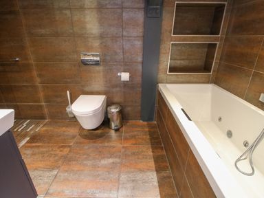 A bathroom with a wall-mounted toilet a bathtub with jets open shelves and a trash bin at Timberdyne in Brimfield near Woofferton