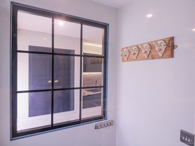 A room with a black framed window looking into a kitchen with purple cabinets and a wall mounted wooden rack with four ceramic masks at Number 1 in Clare