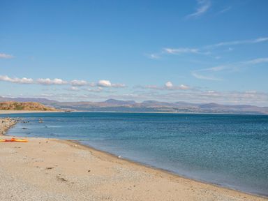 A beach with sand and pebbles next to calm water and distant hills under a blue sky at Gwynfa in Barmouth