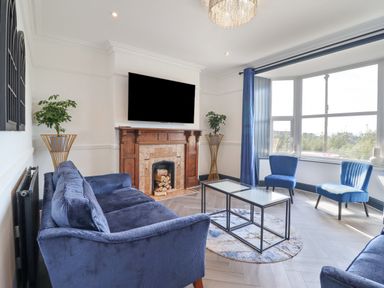 A living room with blue couches and chairs a fireplace with TV above and large window at Gwynfa in Barmouth