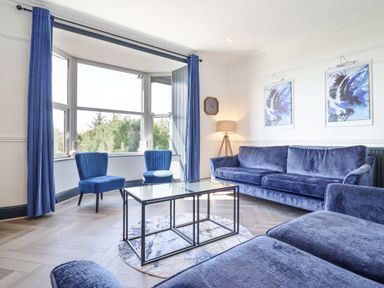 A living room with blue chairs blue curtains a blue sofa a glass coffee table and two paintings on the wall at Gwynfa in Barmouth
