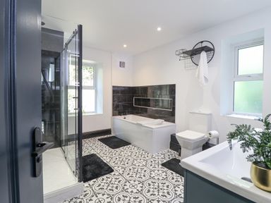 A bathroom with a shower, bathtub, toilet, sink, patterned floor tiles, and two windows at Gwynfa in Barmouth