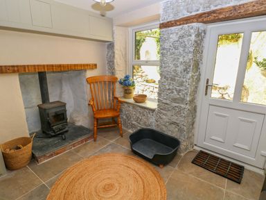 A room with a wood stove next to a wooden chair a basket with firewood a round rug a dog bed and a window and door with stone walls at Suffolk House in Narberth