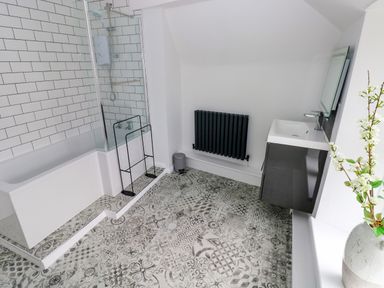 A bathroom with a bathtub shower tiled wall patterned floor black radiator sink and mirror with flowers in a vase at Suffolk House in Narberth