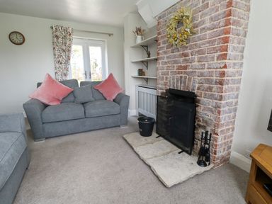 A living room with a grey sofa with pink cushions a brick fireplace and a window with floral curtains at Rempstone Farmhouse in Corfe Castle