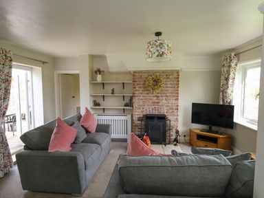 A living room with two gray sofas with pink cushions a brick fireplace a TV on a wooden stand shelves and floral curtains at Rempstone Farmhouse in Corfe Castle
