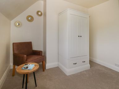 A room corner with a brown chair next to a small wooden table with books and a white wardrobe on a beige carpet at Rempstone Farmhouse in Corfe Castle