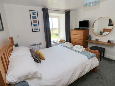A bedroom with a wooden bed white bedding pillows a window with curtains a dresser with a TV a round mirror and a small desk at Grassholm View in Solva