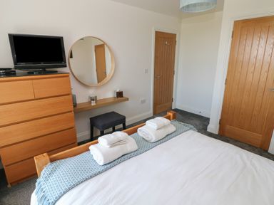 A bedroom with a wooden bed with towels on the blanket a wooden dresser with a television a round wall mirror a small stool and two wooden doors at Grassholm View in Solva