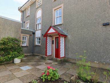 An outdoor entrance to a building with a small porch and red door surrounded by plants and paved courtyard at Grassholm View in Solva