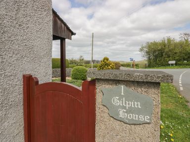 A red wooden gate next to a stone pillar with a sign reading 1 Gilpin House in Levens
