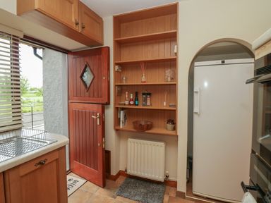 A kitchen with a wooden door open shelves above a radiator a white refrigerator and a window with blinds at 1 Gilpin House Levens