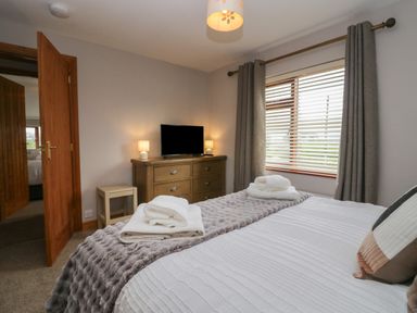 A bedroom with a bed covered in white linens and a gray throw a wooden chest of drawers with a TV two lamps and folded towels on the bed a window with blinds and gray curtains 1 Gilpin House Levens
