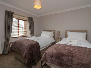 A bedroom with two single beds with white linens and brown blankets a bedside table and window with curtains at 1 Gilpin House in Levens