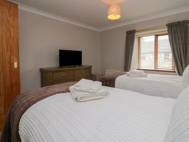 A bedroom with two beds with white linens a wooden dresser with a television and a window with blinds and curtains at 1 Gilpin House in Levens