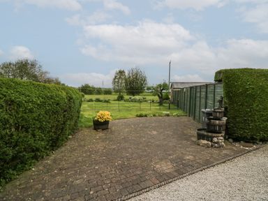An outdoor space with a paved area bordered by green hedges and a green wooden fence with a grassy field and trees beyond at 1 Gilpin House in Levens